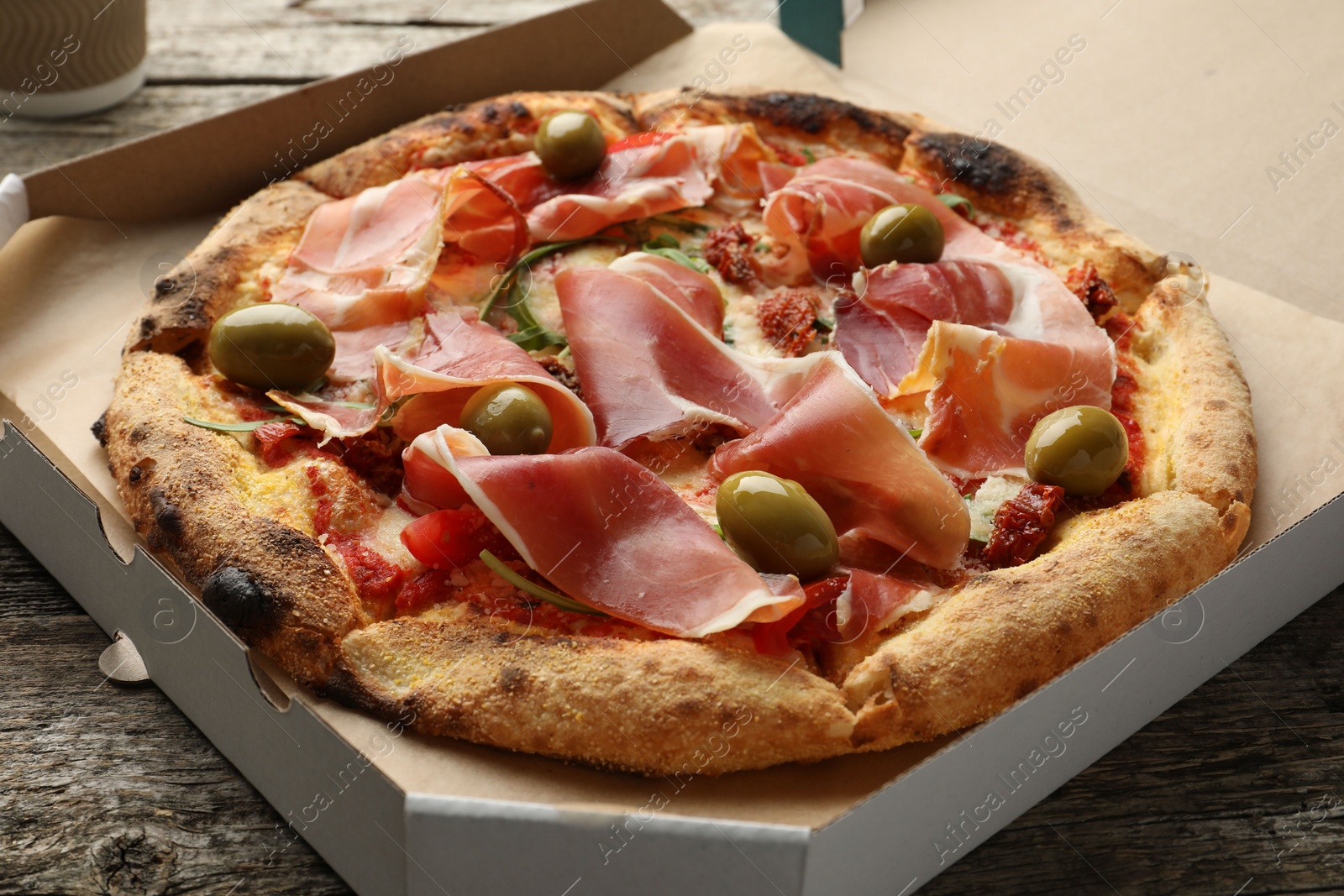 Photo of Tasty pizza with cured ham, olives and sun-dried tomato in cardboard box on wooden table