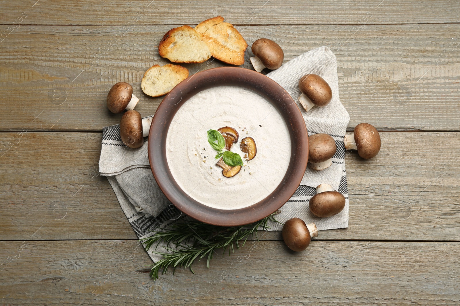 Photo of Tasty homemade mushroom soup in ceramic pot, croutons, fresh champignons and rosemary on wooden table, flat lay