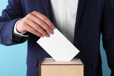 Man putting his vote into ballot box on light blue background, closeup