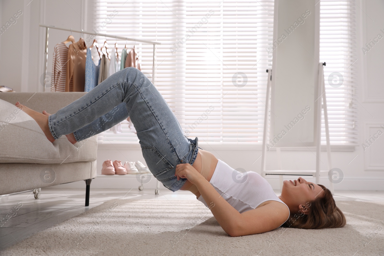 Photo of Young woman struggling to squeeze into tight jeans while lying on floor at home