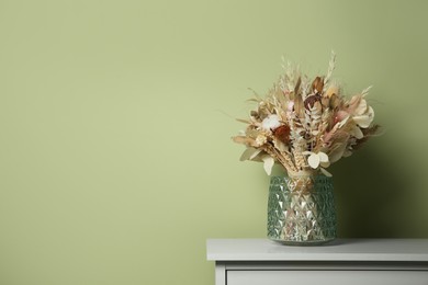 Photo of Beautiful dried flower bouquet in glass vase on white table near olive wall. Space for text