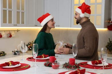 Photo of Happy young man in Santa hat presenting Christmas gift to his girlfriend at table in kitchen