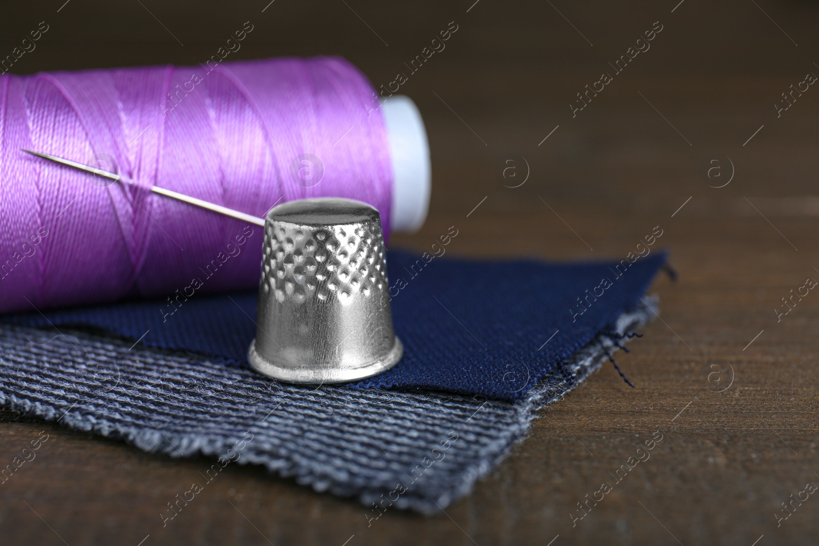 Photo of Silver thimble, thread and needle on wooden table, space for text. Sewing accessories