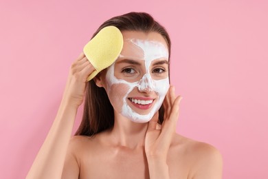 Photo of Happy young woman washing off face mask with sponge on pink background