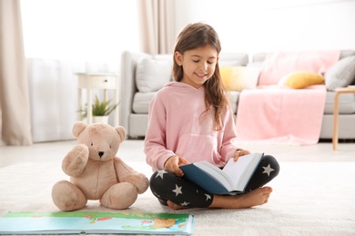 Photo of Cute little girl with teddy bear reading book on floor at home