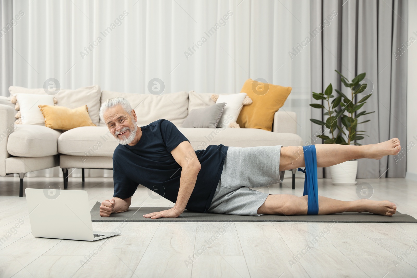 Photo of Senior man doing exercise with fitness elastic band near laptop on mat at home