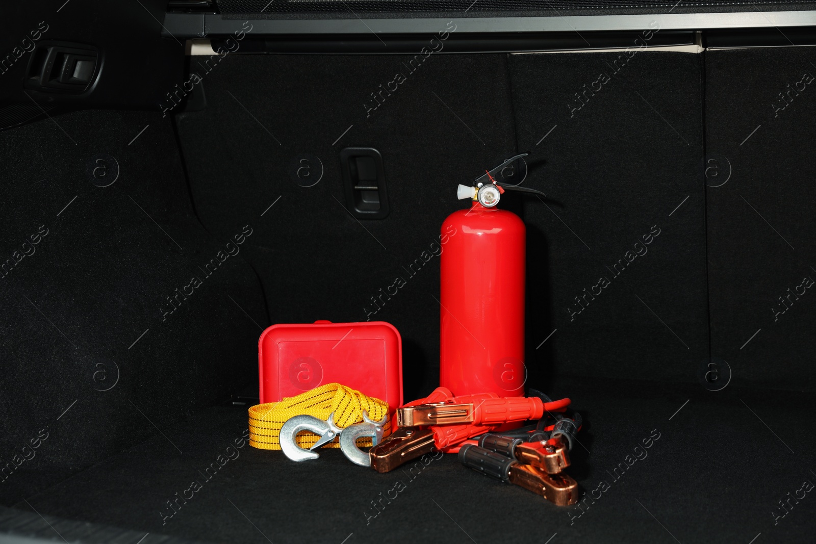 Photo of Red fire extinguisher, battery jumper cables, towing strap and first aid kit in trunk. Car safety