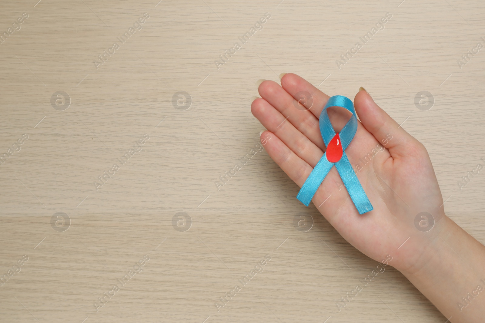 Photo of Woman holding light blue ribbon with paper blood drop at wooden table, top view and space for text. Diabetes awareness