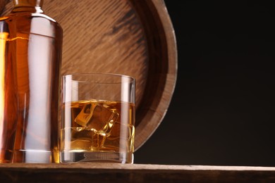 Photo of Whiskey with ice cubes in glass, bottle and barrel on wooden table against black background, space for text