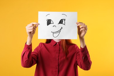 Woman hiding behind sheet of paper with happy face on yellow background