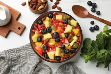 Delicious fruit salad in bowl, berries, fresh mint and nuts on white marble table, flat lay