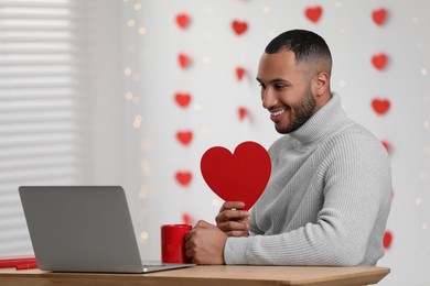 Photo of Valentine's day celebration in long distance relationship. Man holding red paper heart while having video chat with his girlfriend via laptop indoors