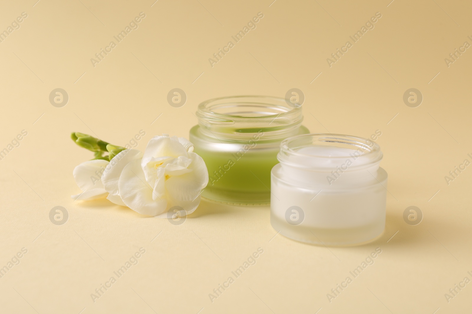 Photo of Moisturizing cream in open jars and freesia flower on beige background. Body care products