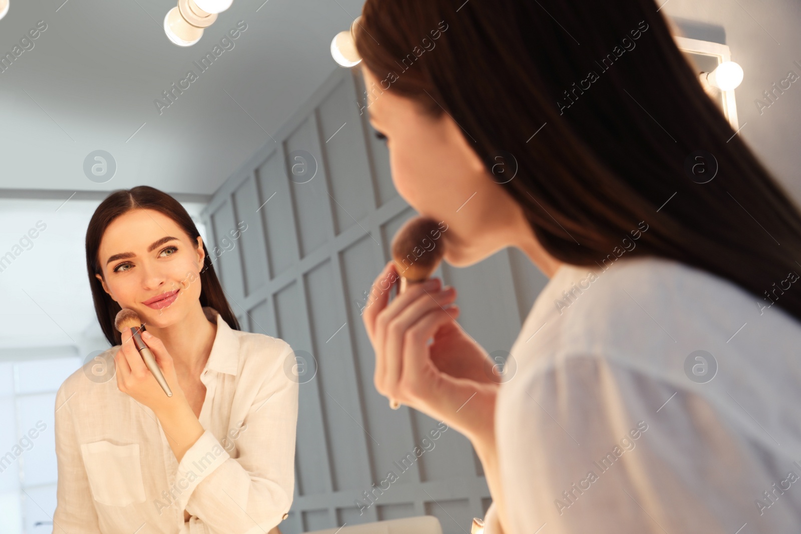 Photo of Young woman applying make up near illuminated mirror indoors