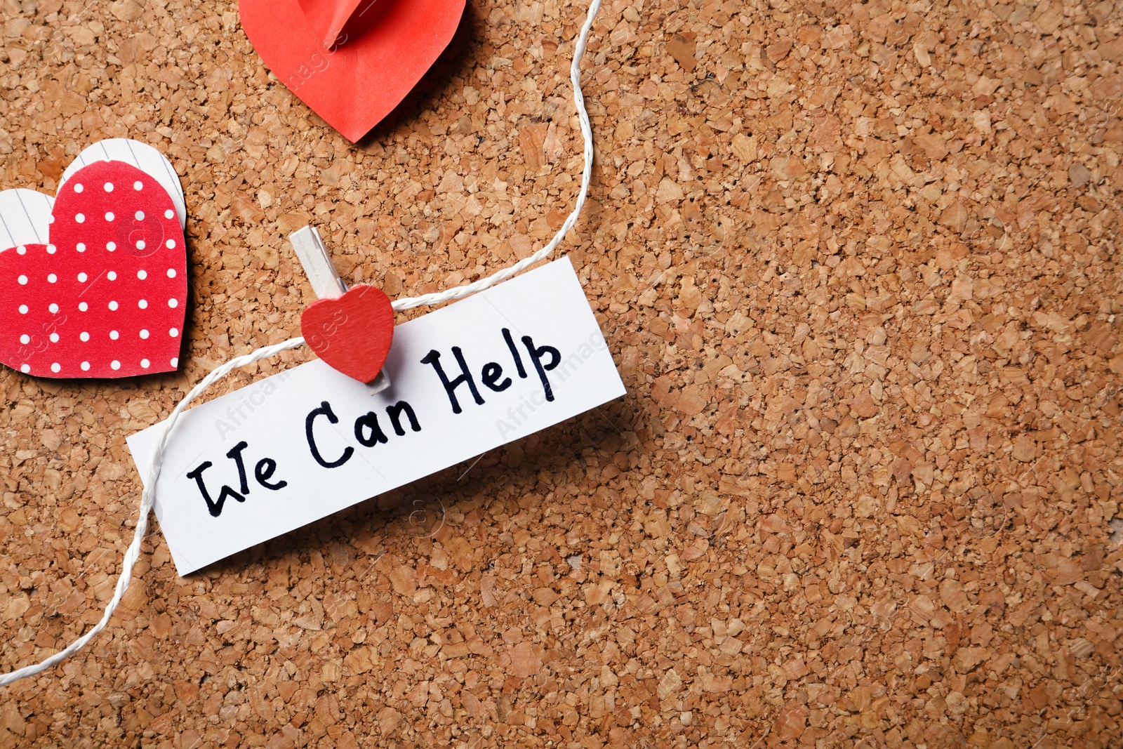 Photo of Paper hearts and note with words WE CAN HELP attached to string on cork board. Space for text