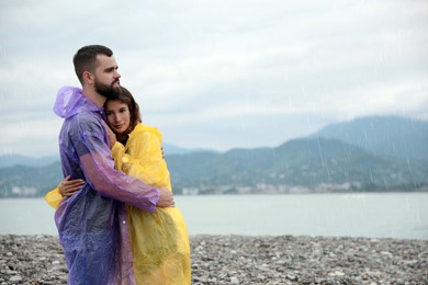 Young couple in raincoats enjoying time together under rain on beach, space for text