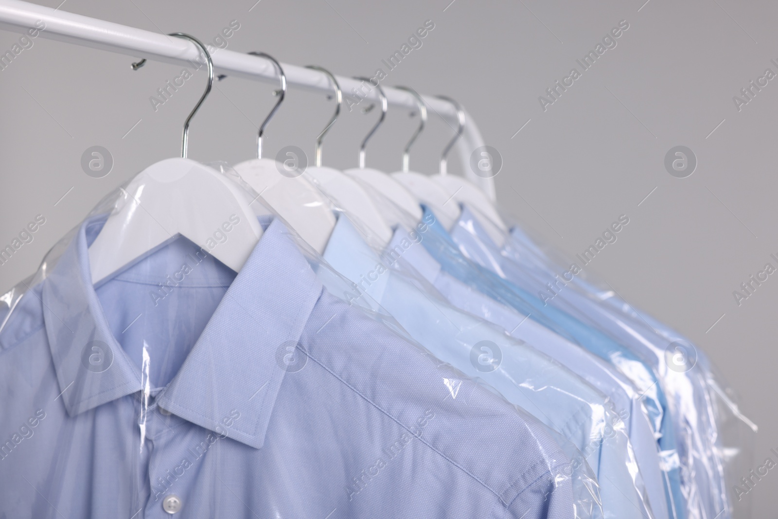 Photo of Dry-cleaning service. Many different clothes in plastic bags hanging on rack against grey background, closeup