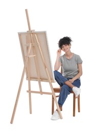 Young woman holding brush near easel with canvas against white background