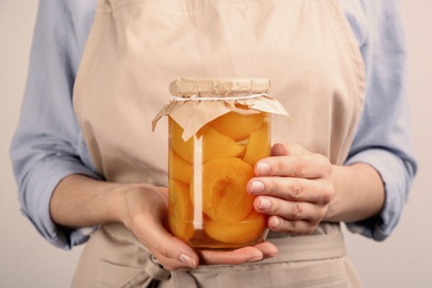 Woman holding glass jar of pickled peaches on light background, closeup