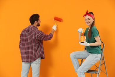 Photo of Man painting orange wall and happy woman holding can of dye with brush. Interior design