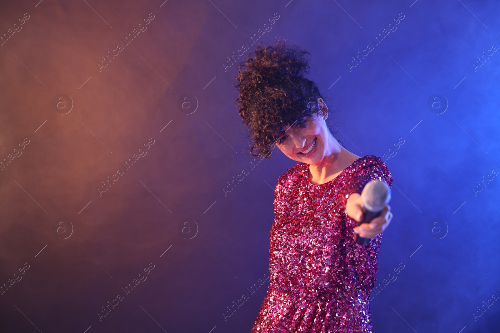 Photo of Beautiful young woman with microphone on color background in neon lights and smoke