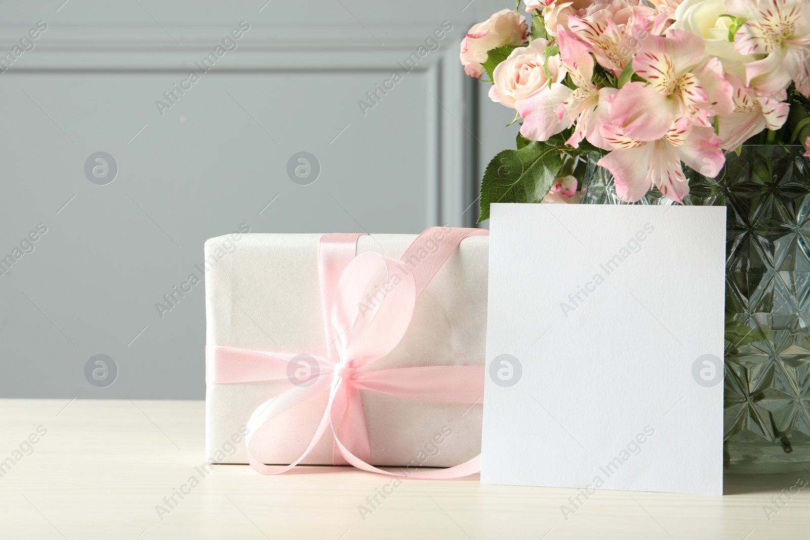 Photo of Happy Mother's Day. Gift box, blank card and bouquet of beautiful flowers in vase on white table near grey wall