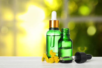 Bottles of essential oil and flowers on white wooden table against blurred background