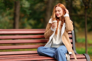 Photo of Beautiful woman with paper cup sitting on bench in autumn park. Space for text