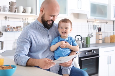 Photo of Dad reading newspaper while holding little son in kitchen