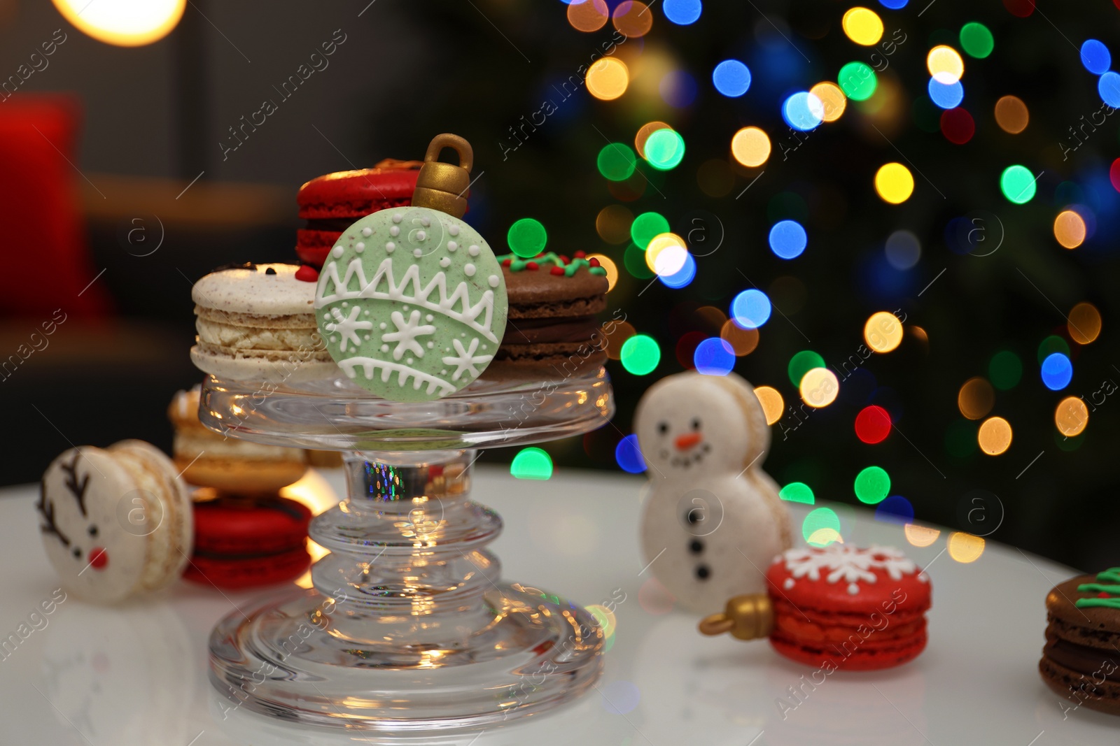 Photo of Beautifully decorated Christmas macarons on white table against blurred festive lights, space for text