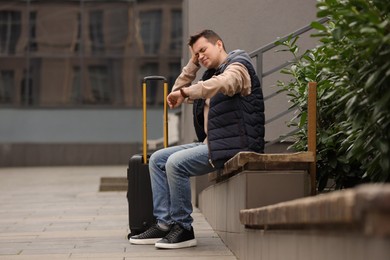 Being late. Worried man with suitcase looking at watch on bench outdoors, space for text