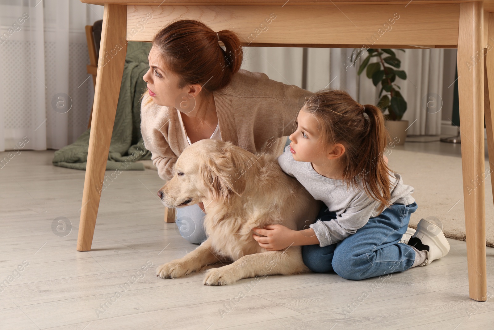 Photo of Scared mother with her little daughter and dog hiding under table in living room during earthquake