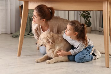 Photo of Scared mother with her little daughter and dog hiding under table in living room during earthquake