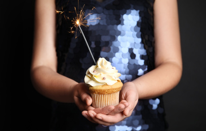 Photo of Woman holding birthday cupcake with sparkler on black background, closeup