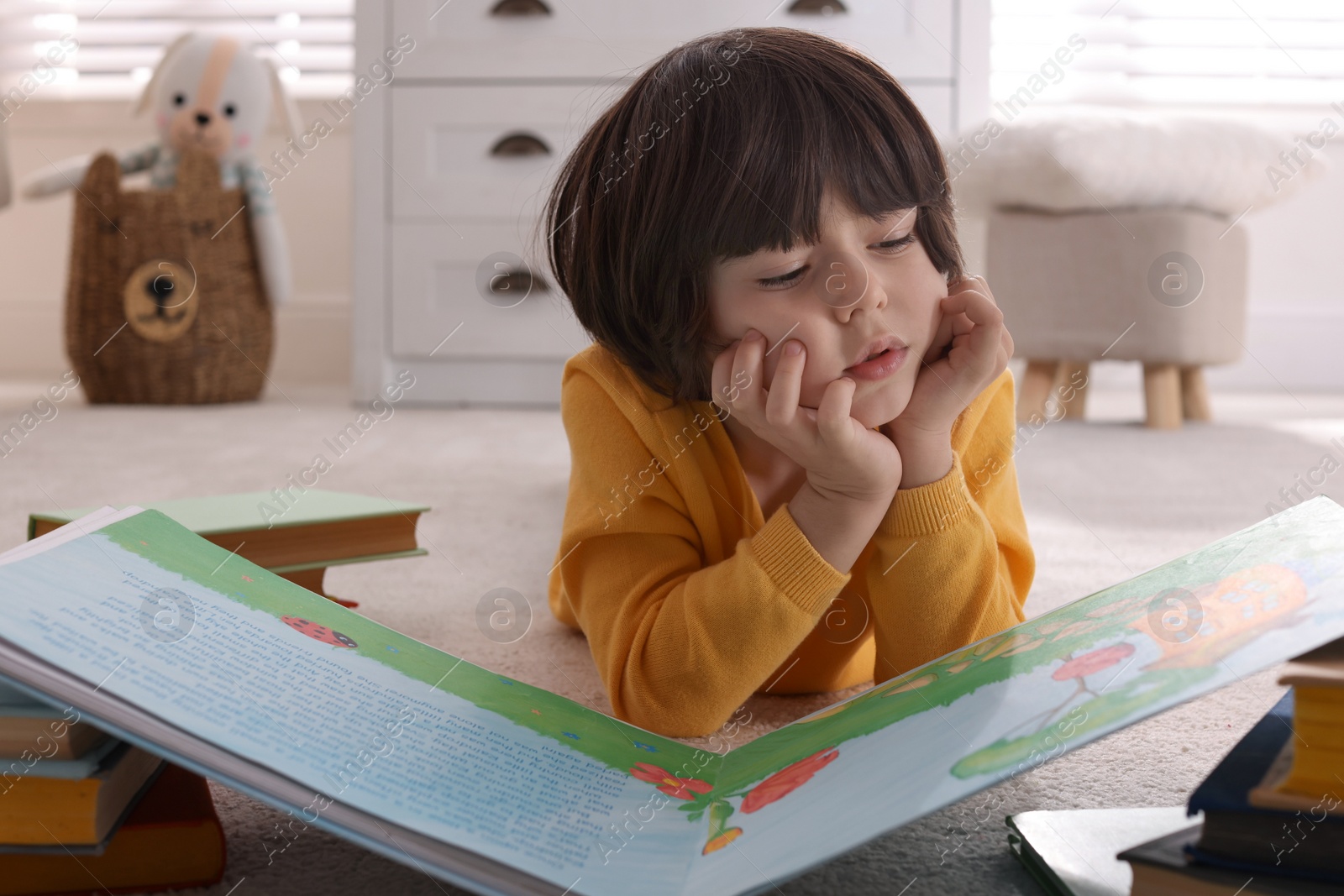 Photo of Cute little boy reading book on floor at home