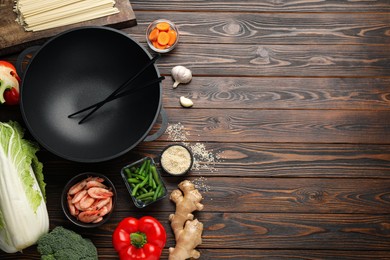 Photo of Empty iron wok with chopsticks and raw ingredients on wooden table, flat lay. Space for text