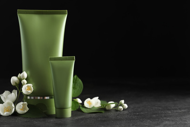 Photo of Tubes of cosmetic products and flowers on grey stone table against black background. Space for text