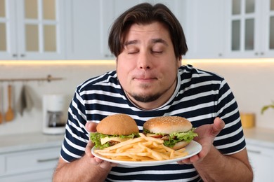 Photo of Happy overweight man holding plate with tasty burgers and French fries in kitchen