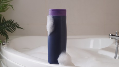 Photo of Bottle of bubble bath with foam on tub indoors