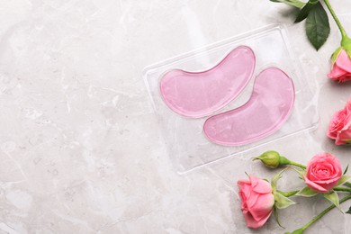 Photo of Package of under eye patches and rose flowers on light grey table, flat lay with space for text. Cosmetic product