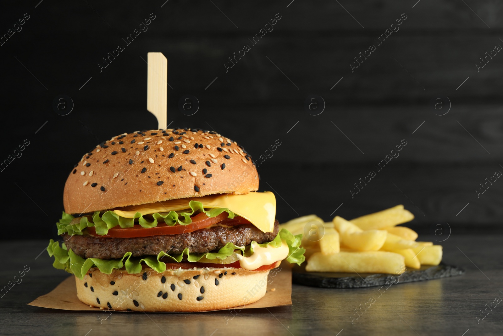 Burger with delicious patty and french fries on gray table against dark background, space for text Photo of Burger with delicious patty and french fries on gray table against dark background, space for text