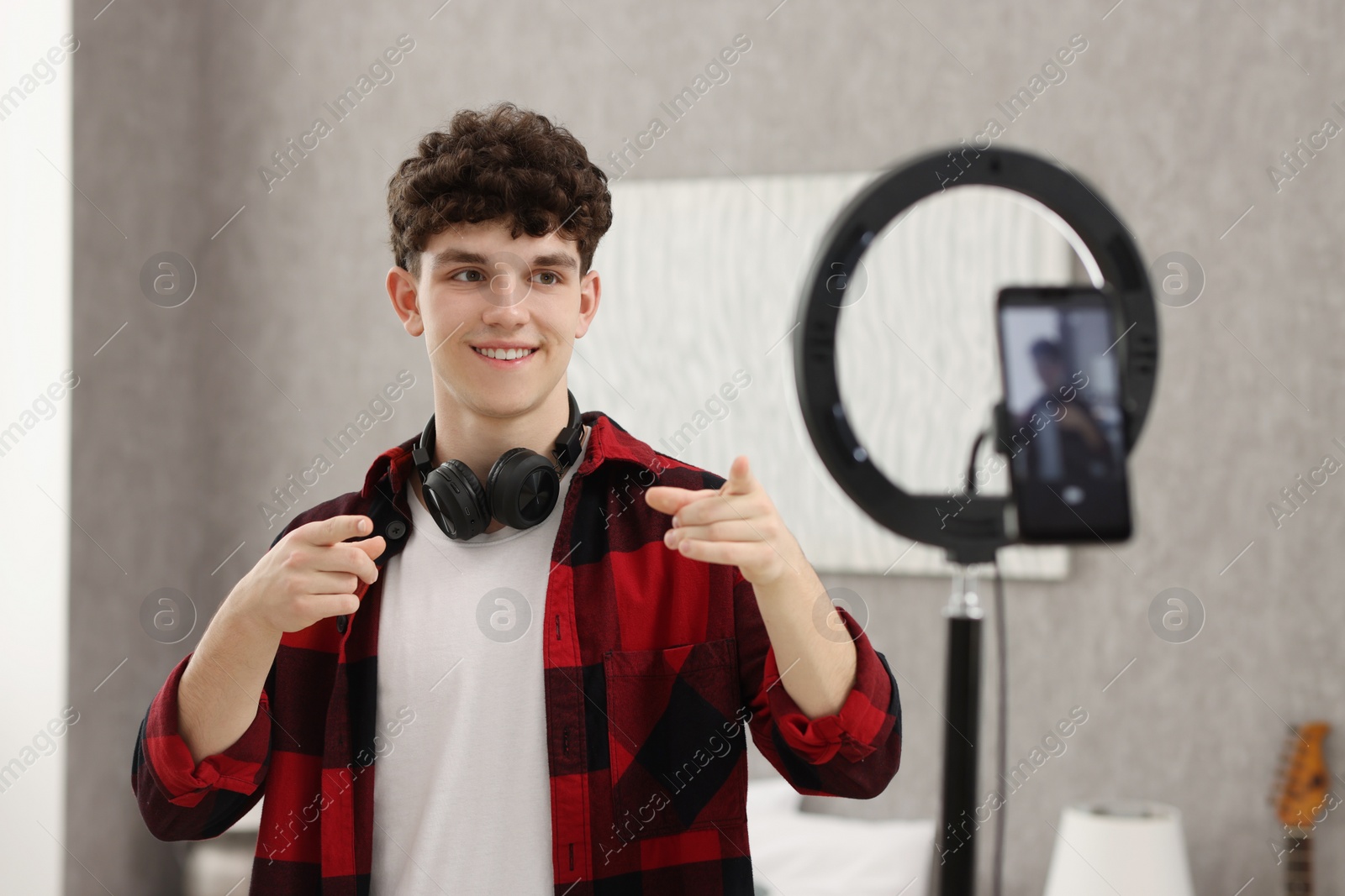 Photo of Smiling teenage blogger pointing at his smartphone while streaming at home