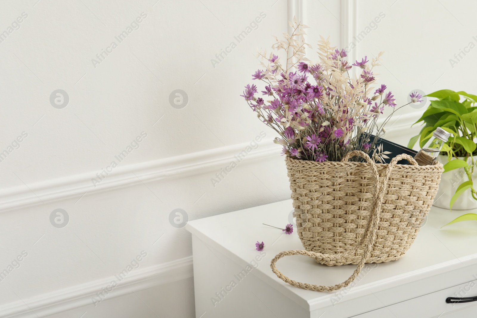 Photo of Stylish beach bag with beautiful wildflowers, bottle of water and book on cabinet in room. Space for text