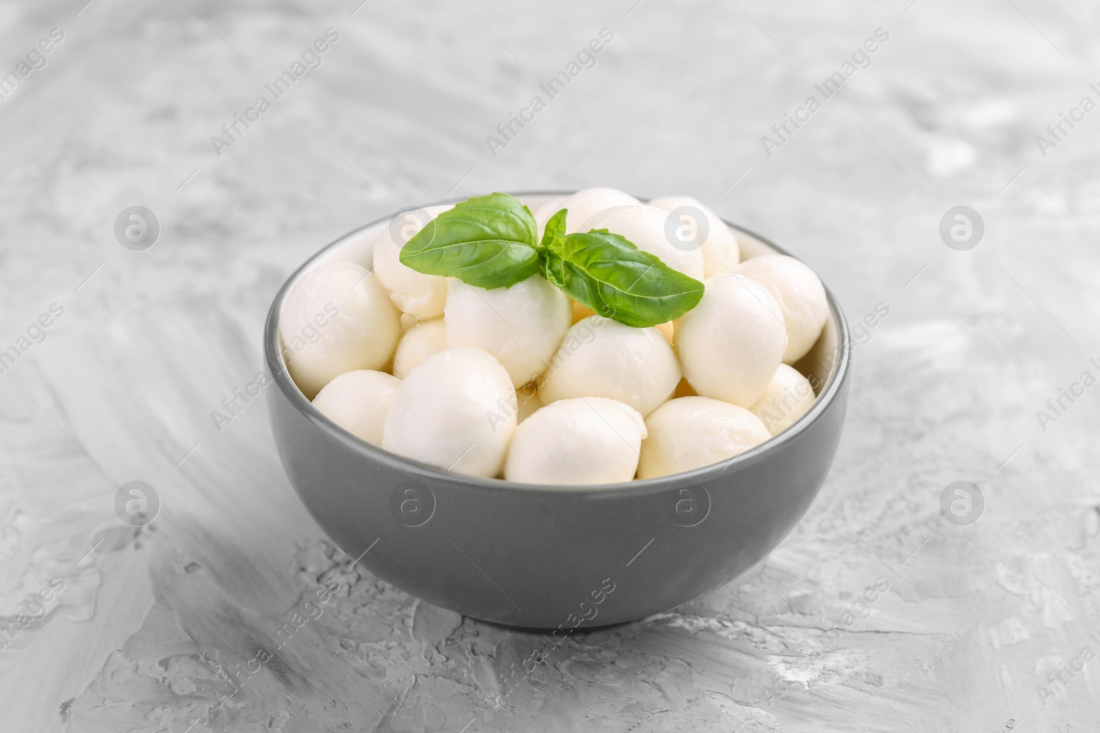 Photo of Tasty mozzarella balls and basil leaves in bowl on grey table, closeup