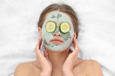 Photo of Beautiful woman with clay facial mask and cucumber slices on white fabric, above view