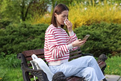 Woman with napkin suffering from seasonal allergy while using smartphone on bench in park