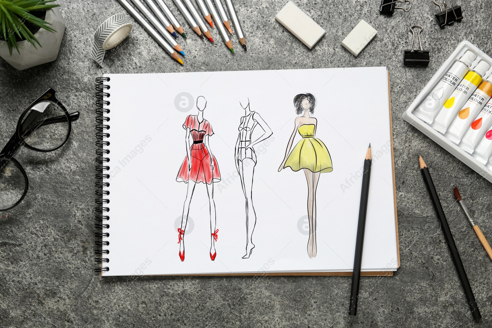 Image of Sketches of different clothes in pad on grey textured table. Fashion designer's desk with stationery, flat lay