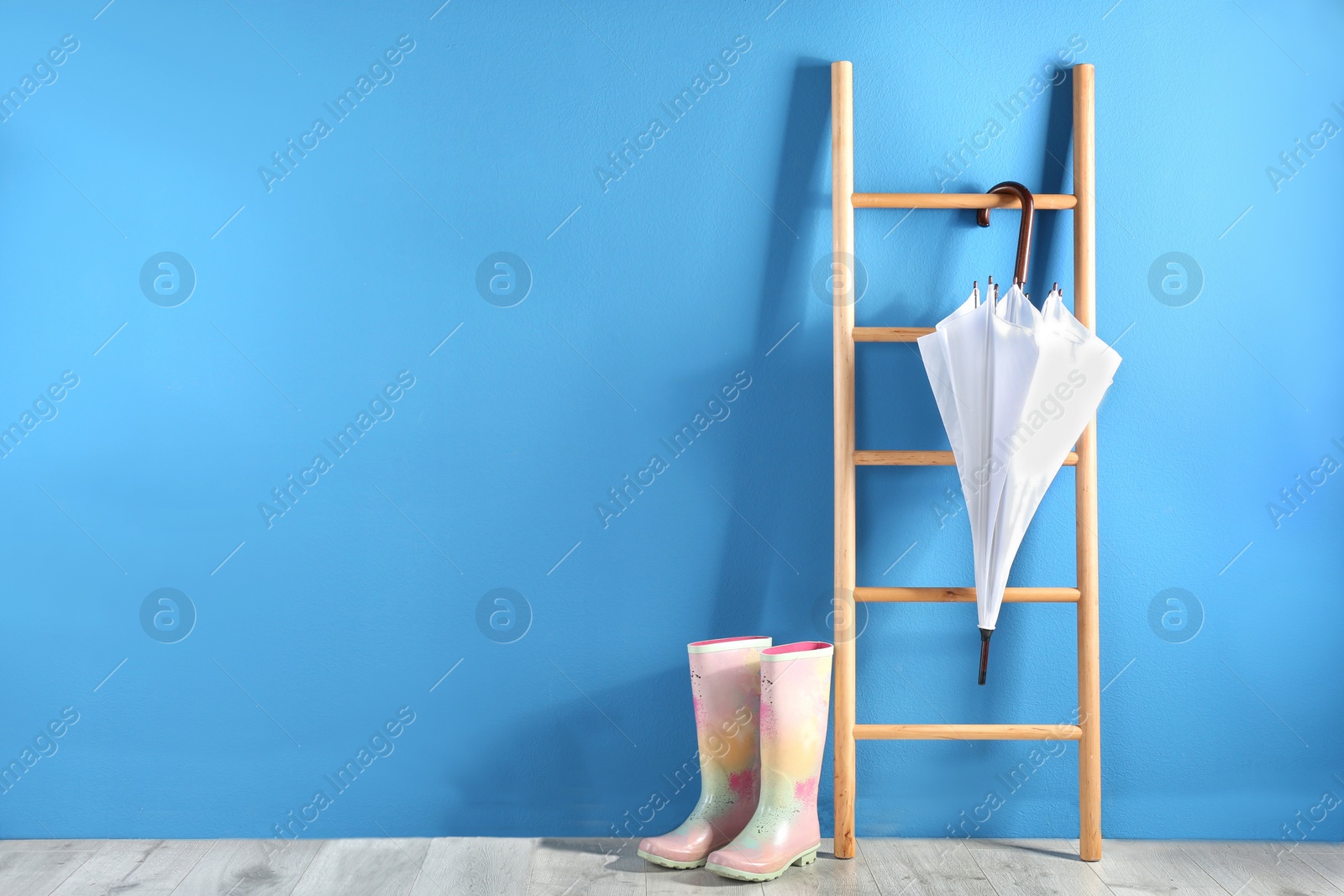 Photo of Beautiful white umbrella on ladder and rubber boots near blue wall. Space for text