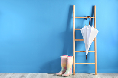 Beautiful white umbrella on ladder and rubber boots near blue wall. Space for text