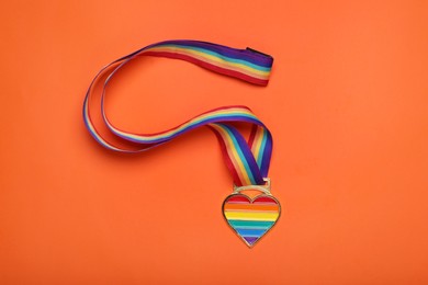 Photo of Rainbow ribbon with heart shaped pendant on orange background, top view. LGBT pride